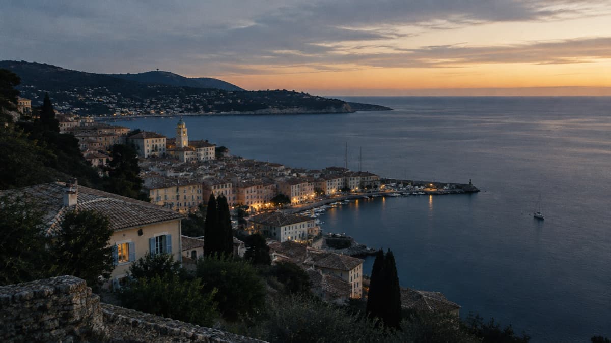 Une ville du littoral méditerranéen vue d’une colline au crépuscule, les premières lumières allumées et la mer sombre au-delà